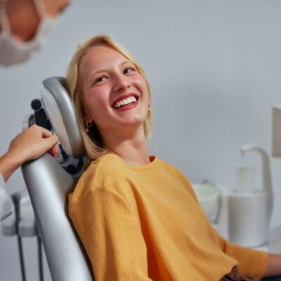 Female dentist wearing medical overalls, mask and gloves talking to young woman lying on dental chair