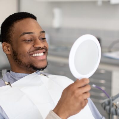 Portrait Of Happy Dental Clinic Patient Looking At Mirror After Treatment, Cheerful African American Guy Sitting In Chair In Stomatological Cabinet And Enjoying His Perfect Smile, Closeup